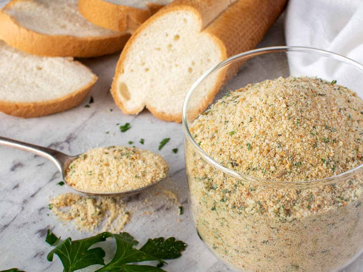 Breadcrumbs in glass jar with a spoonful beside the jar and bread in the background.