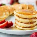 four mini pancakes stacked on a white plate with syrup dripping off and more pancakes and strawberries around.
