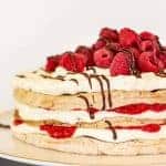 image with text. text reads decadent and elegant raspberry torte. image is three layer meringue cake with cream, raspberries and chocolate on a white cake stand with raspberries scattered underneath.