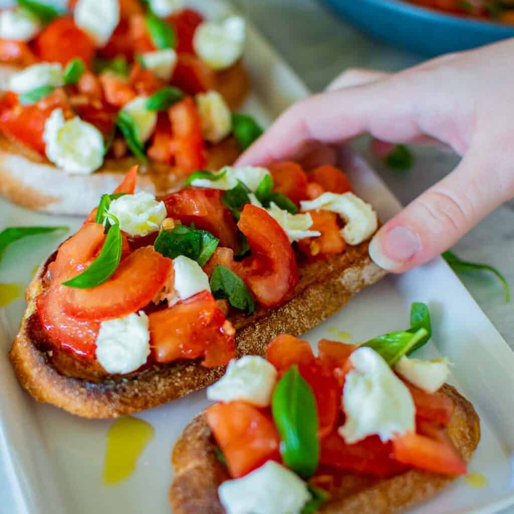 A hand reaching for toasted slice of bread topped with chopped tomato and mozzarella.