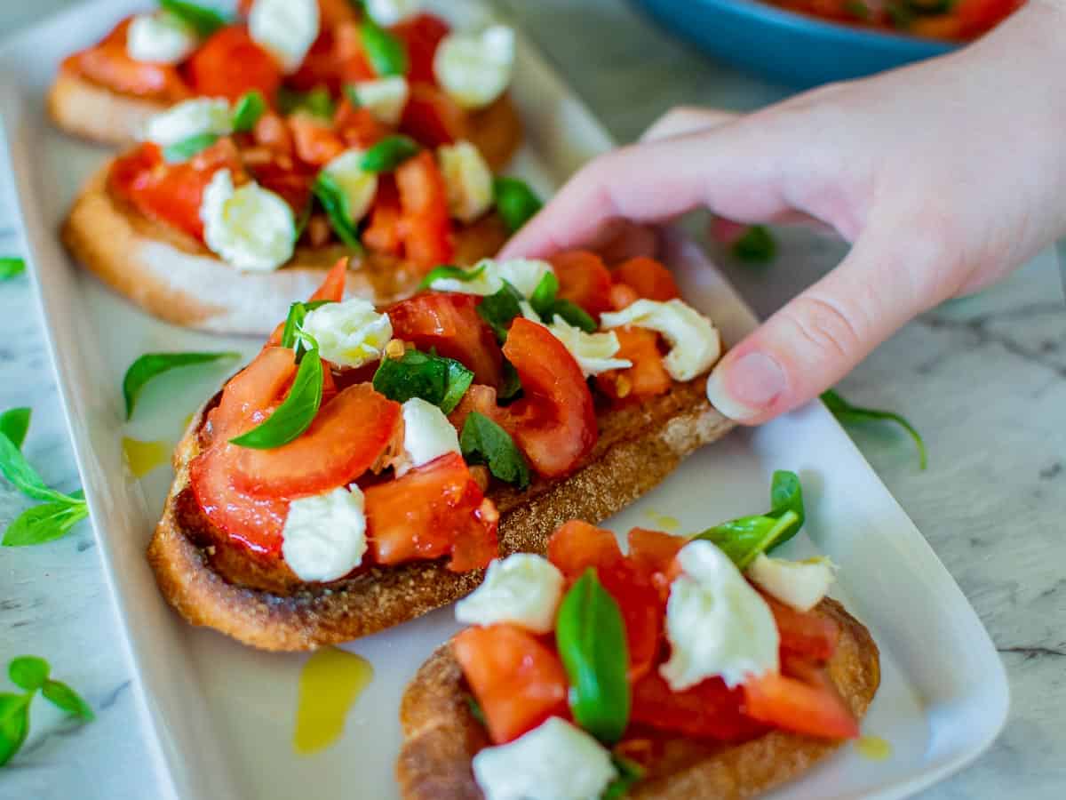 A hand reaching for toasted slice of bread topped with chopped tomato and mozzarella.