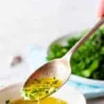 image with text. text reads "Easy and delicious garlic butter sauce". Image is A silver teaspoonful of garlic butter sauce taken from a small white bowl with garlic and broccolini in the background.