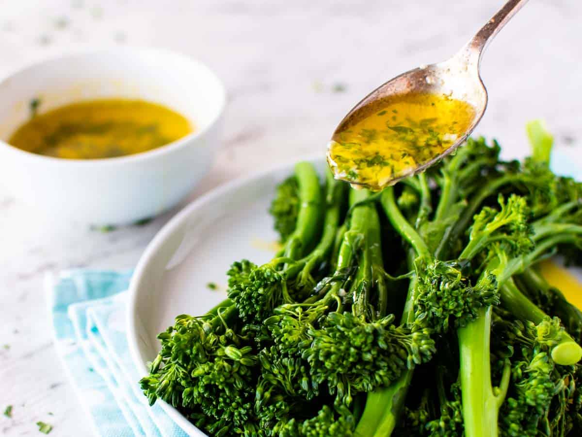 A teaspoon of the sauce being spooned over broccolini with a white bowl in the background.