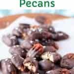 image with text. text reads "salted chocolate covered pecans". image is Pile of chocolate dipped pecans sprinkled with flaked salt and resting on white parchment paper.