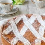 image with text. text reads "Italian wheat berry pie Pastiera Napoletana". Image is round high crusted pie with lattice top dusted with powdered sugar, espresso in the background.