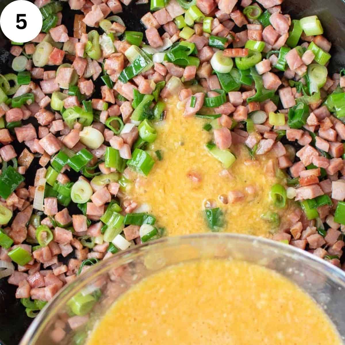 Beaten eggs being poured over diced ham and chopped green onions.