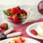 Coconut panna cotta on a white plate topped with sliced strawberries and passion fruit with more in the background.