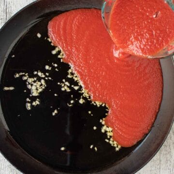 Tomato puree being poured into a black skillet containing oil and garlic.