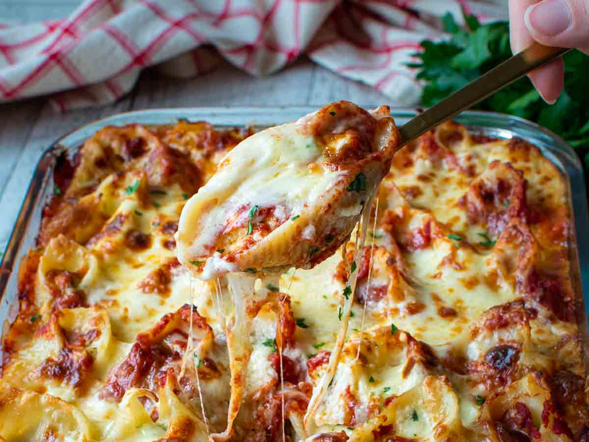 A filled pasta shell being lifted from a pan full of pasta shells with cheesy string pulls.