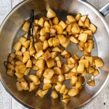 Cooked chopped apple with ground cinnamon and a split vanilla bean in a frying pan.