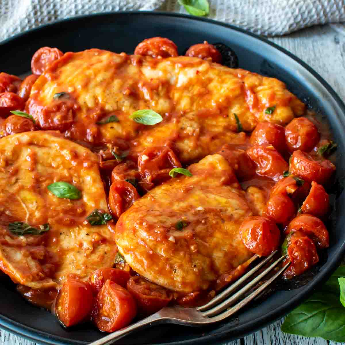 Chicken pomodoro with cherry tomatoes and fresh basil scattered around on a black plate.