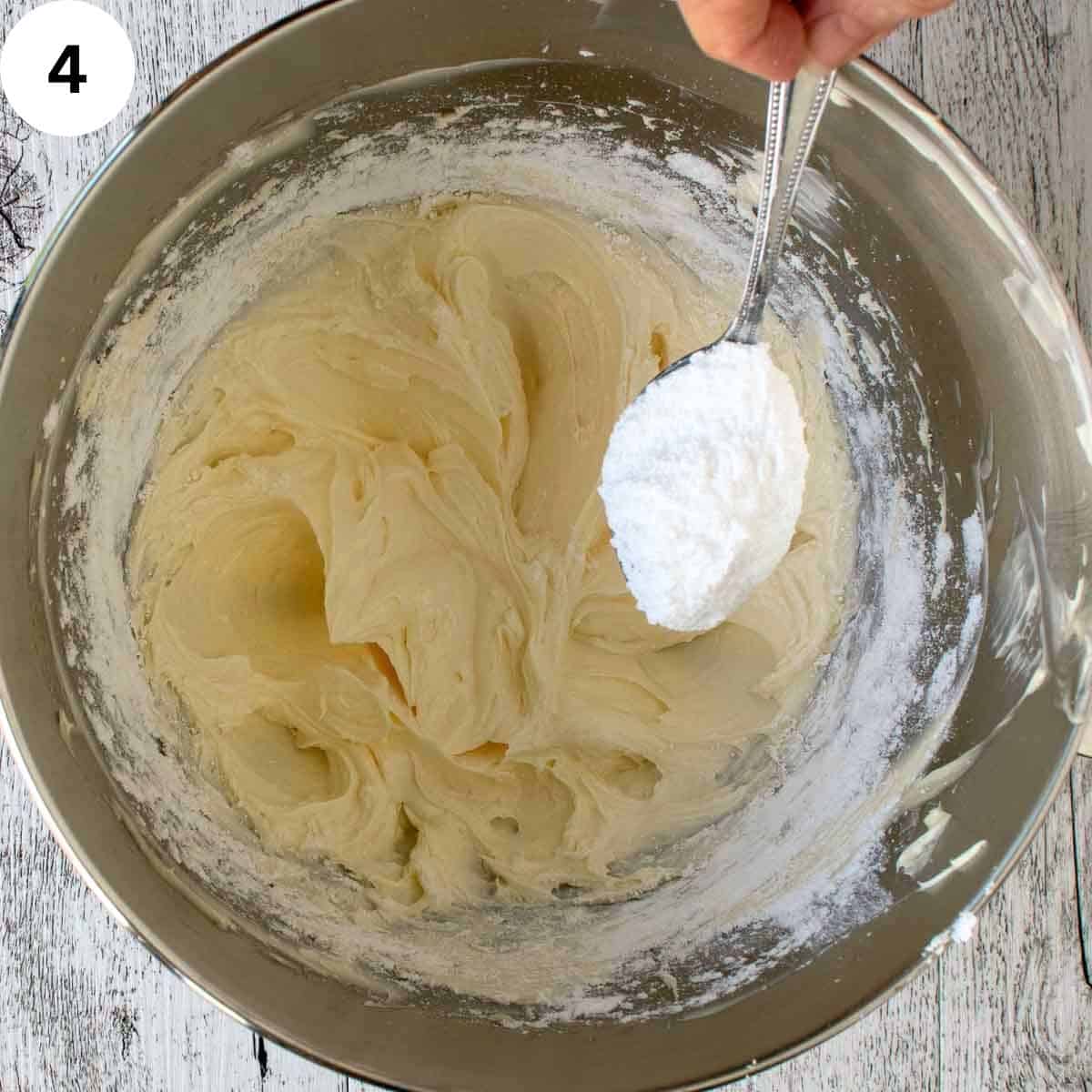 Powdered sugar being spooned into bowl containing creamy butter mixture.