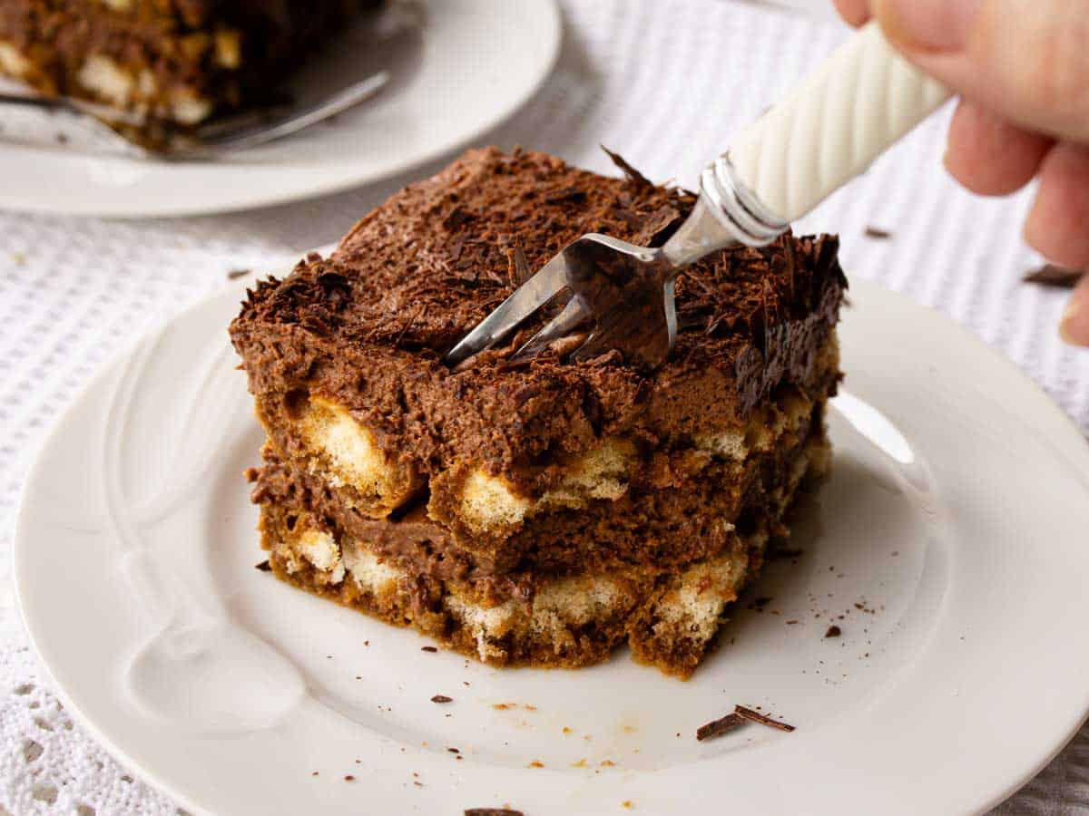 Square of tiramisu with chocolate on a white plate with a fork plunged in.