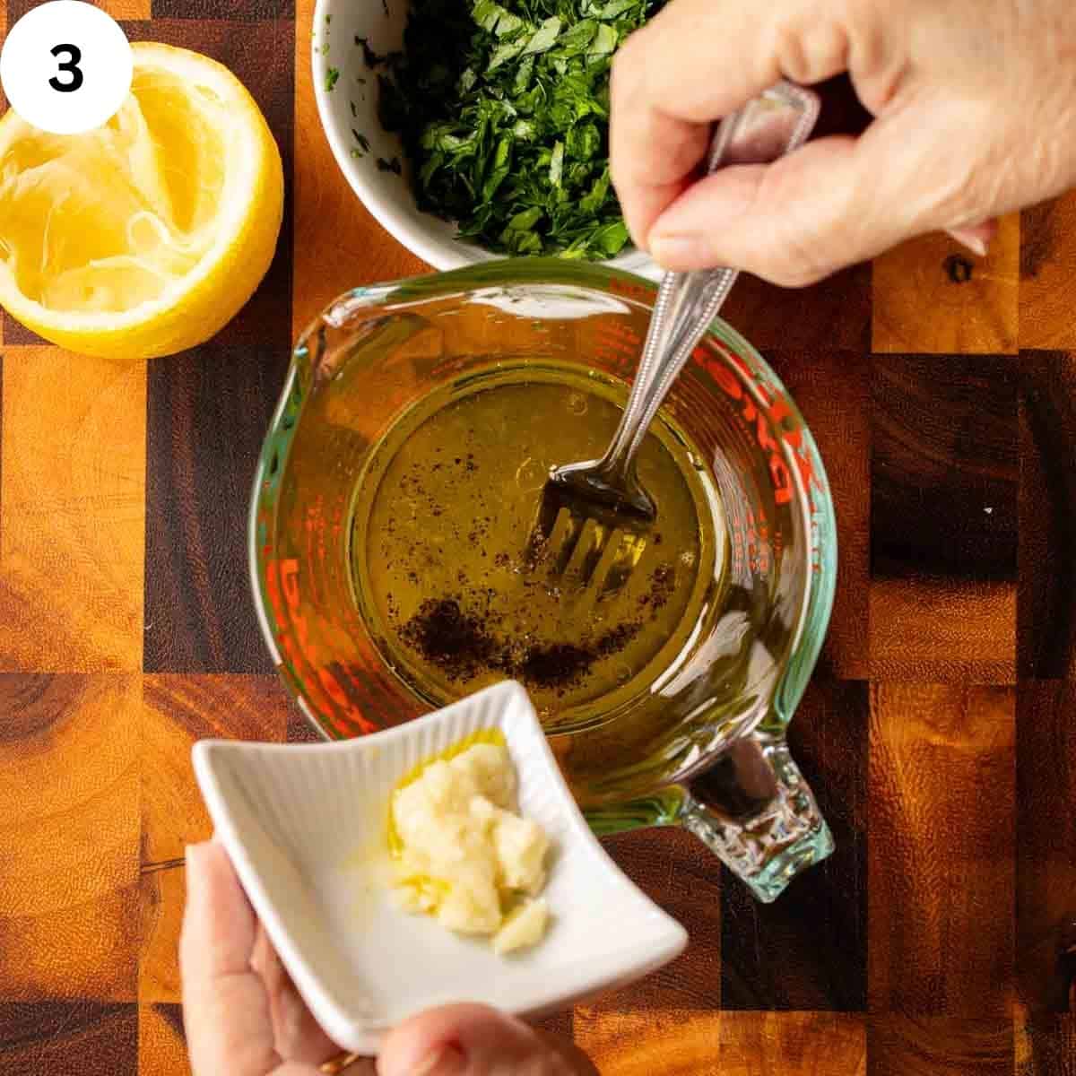 A fork in a glass jug of oil with garlic being added; parsley and lemon on the side.