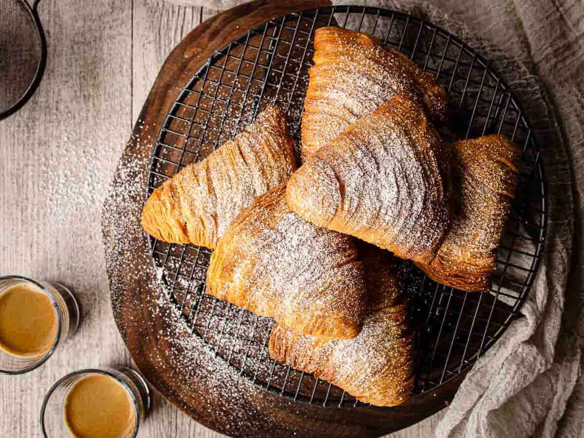 Golden baked clam shaped pastries with many flaky layers on a wire rack viewed from above.