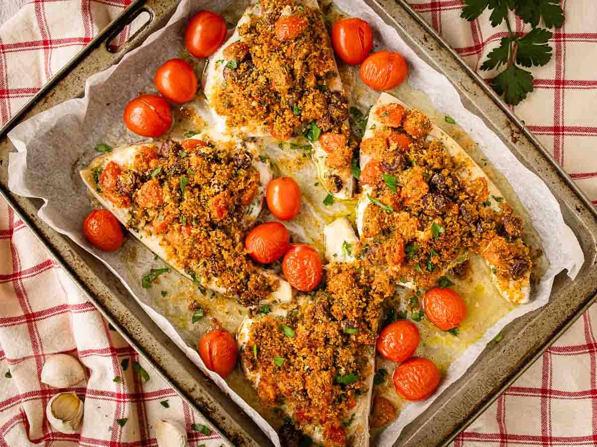 Four breadcrumb topped swordfish steaks in a baking pan surrounded by cherry tomatoes.