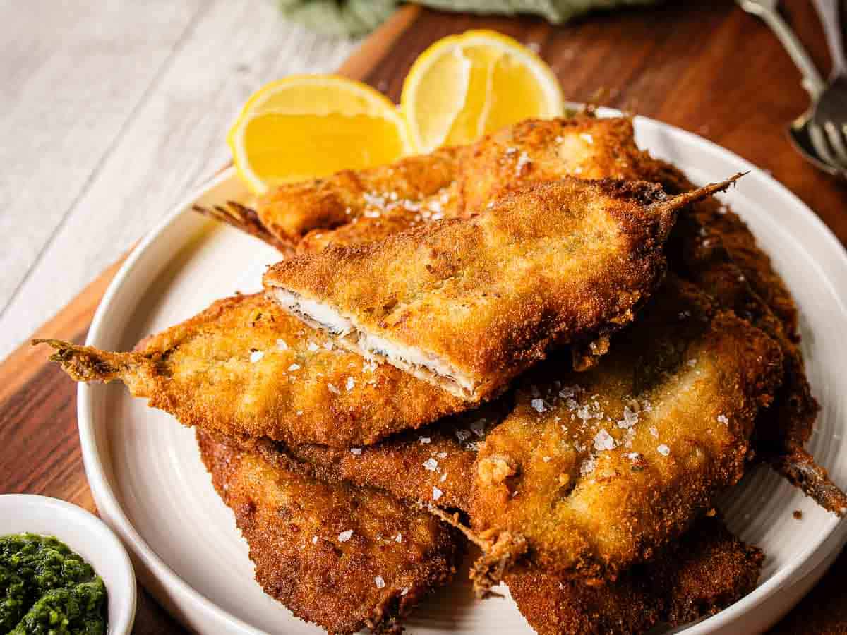 Fried breaded sardines piled on a plate with one cut in half.