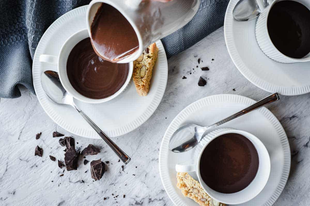 Dark creamy chocolate mixture being poured into a cup with two other cups filled to the side.
