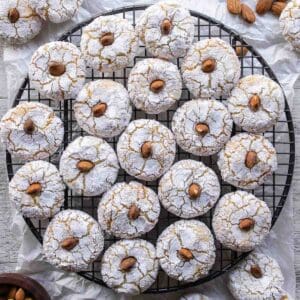 Amaretti Cookies on a round wire rack viewed from above. 3 cups (288 grams) ground almonds 3 egg whites 1 ¼ cup (250 grams) granulated white sugar 1 teaspoon almond extract Powdered sugar Almonds for decorating Prepare two baking sheets by lining them with parchment paper and preheat the oven to 350ºF/180°C. Combine the ground almonds with the sugar in a large bowl. Whip the egg whites until foamy with a fork. Whisk in the almond extract. Then add the egg whites and almond extract mixture. Stir until it becomes too difficult then use your hand to knead into a soft dough. Cover with plastic wrap and rest the mixture for 1 hour. Roll a tablespoon of the mixture into balls. Then roll in granulated sugar followed by powdered sugar. Arrange on a prepared baking sheet. Press down with two fingers to slightly flatten. Insert an almond, if using. Bake in a preheated oven at 350ºF/180°C for about 15 minutes, as they swell and darken remove the soft amaretti from the oven. Allow to cool for 10 minutes before transferring to a wire rack to cool completely. Store in an airtight container.