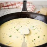 A close-up of a skillet filled with Garlic Parmesan sauce, with a metal spoon resting in it.