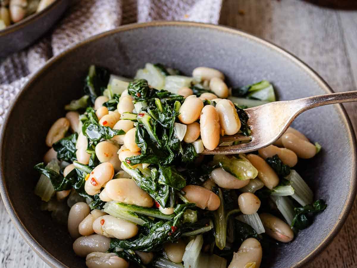 Close up of white beans and cooked greens on a fork with more in a bowl.