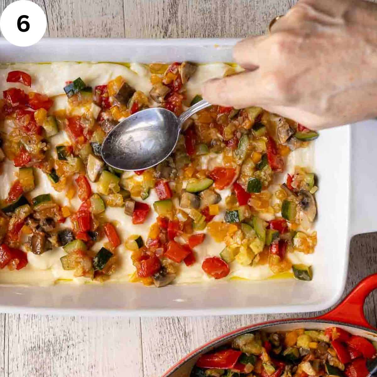 Spreading a mixture of cooked vegetables over white sauce and lasagna noodles in a lasagna pan.