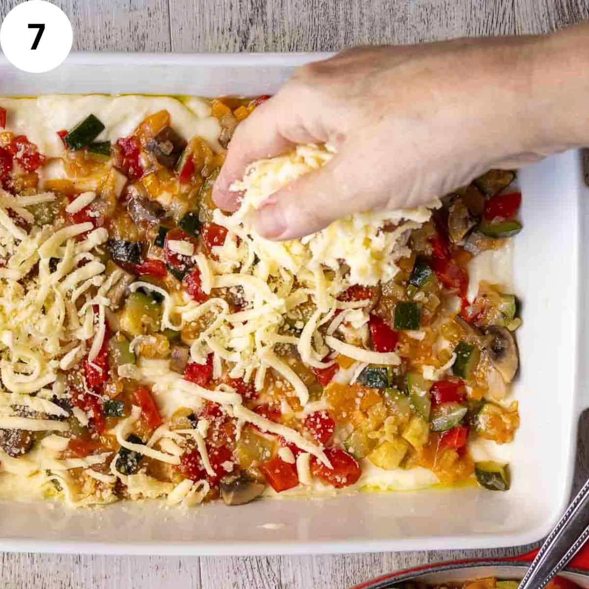 Sprinkling cheese over cooked mixed vegetables in a lasagna dish.