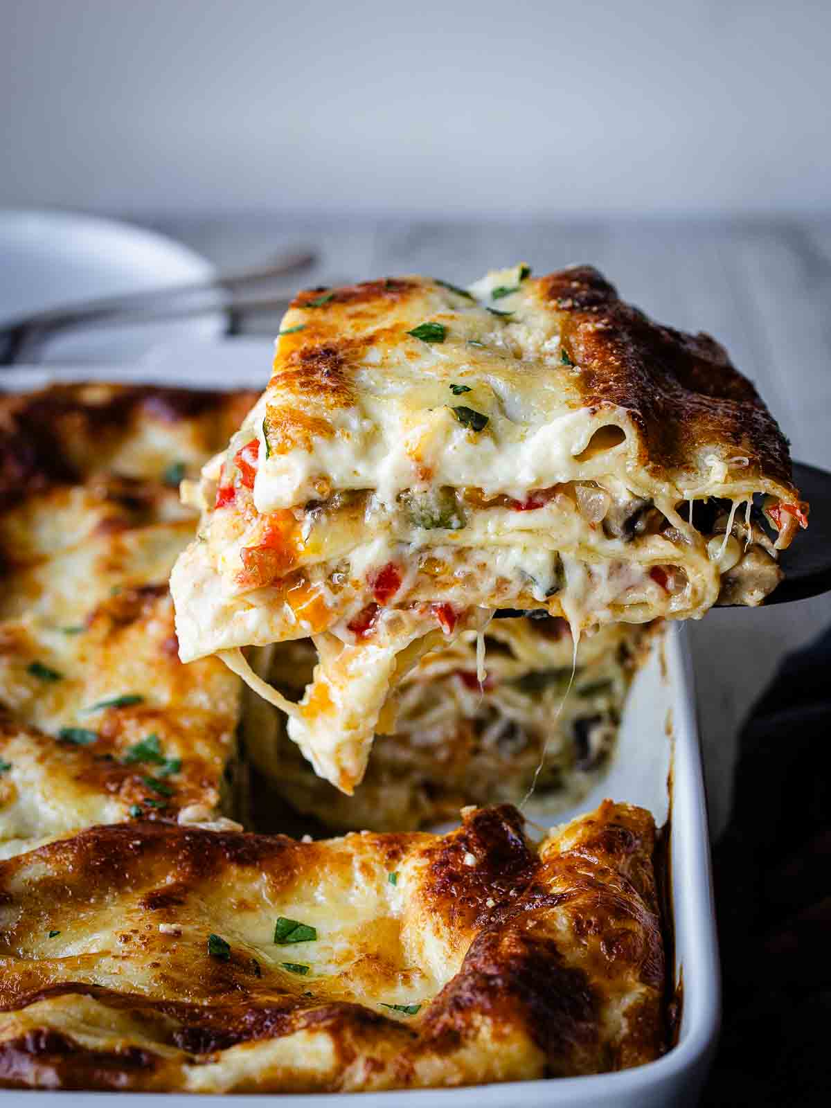 A slice of vegetable lasagna with white sauce being lifted out of a baking dish.