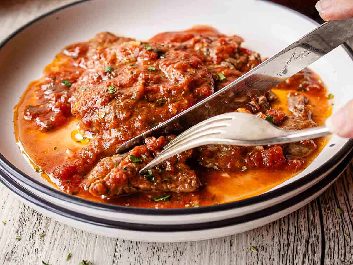 Fork and knife cutting a thin steak smothered in tomato gravy.