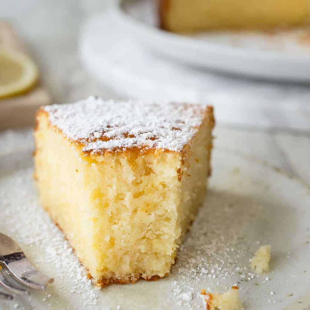 A half eaten wedge of lemon and ricotta cake on a plate with the whole cake in the background.