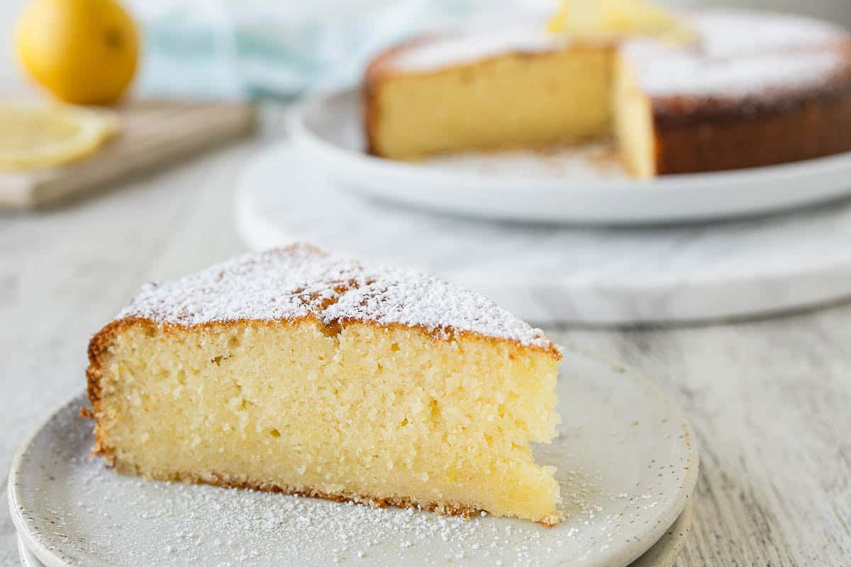Wedge of cake on a plate viewed from the side with a whole cake in the background.