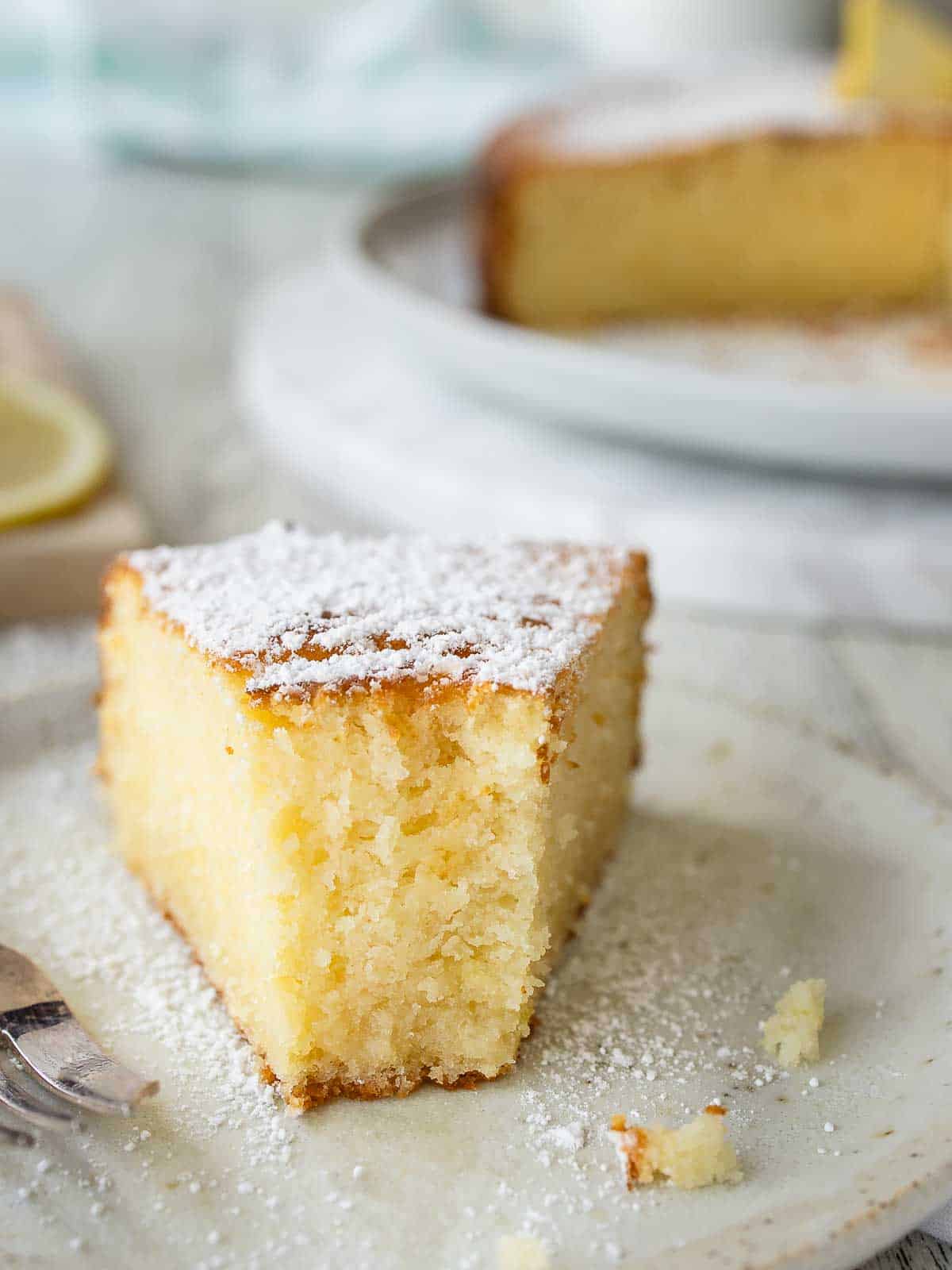 A half eaten wedge of lemon and ricotta cake on a plate with the whole cake in the background.