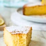 A half eaten wedge of lemon and ricotta cake on a plate with the whole cake in the background.