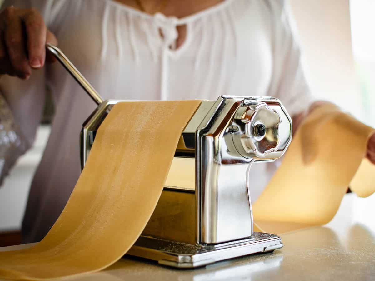 Making fresh pasta a home requires only two ingredients - flour and eggs. Learn how to make your own egg pasta with this step by step recipe. #homemade #homemadepasta #eggpasta #italianrecipe #pastarecipe Woman rolling homemade pasta through a hand cranked pasta machine.