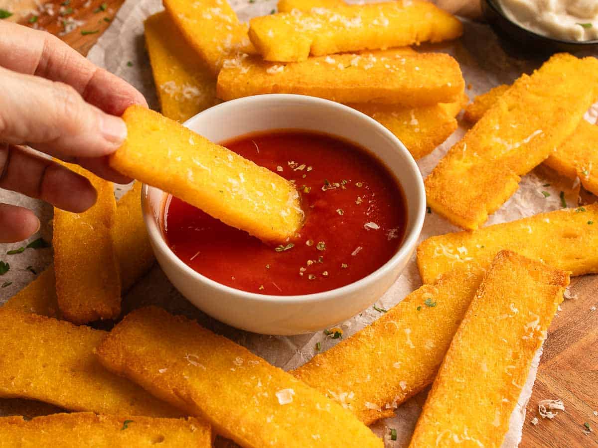 Polenta fries surrounding a bowl of marinara sauce with one fry being dipped into the sauce.