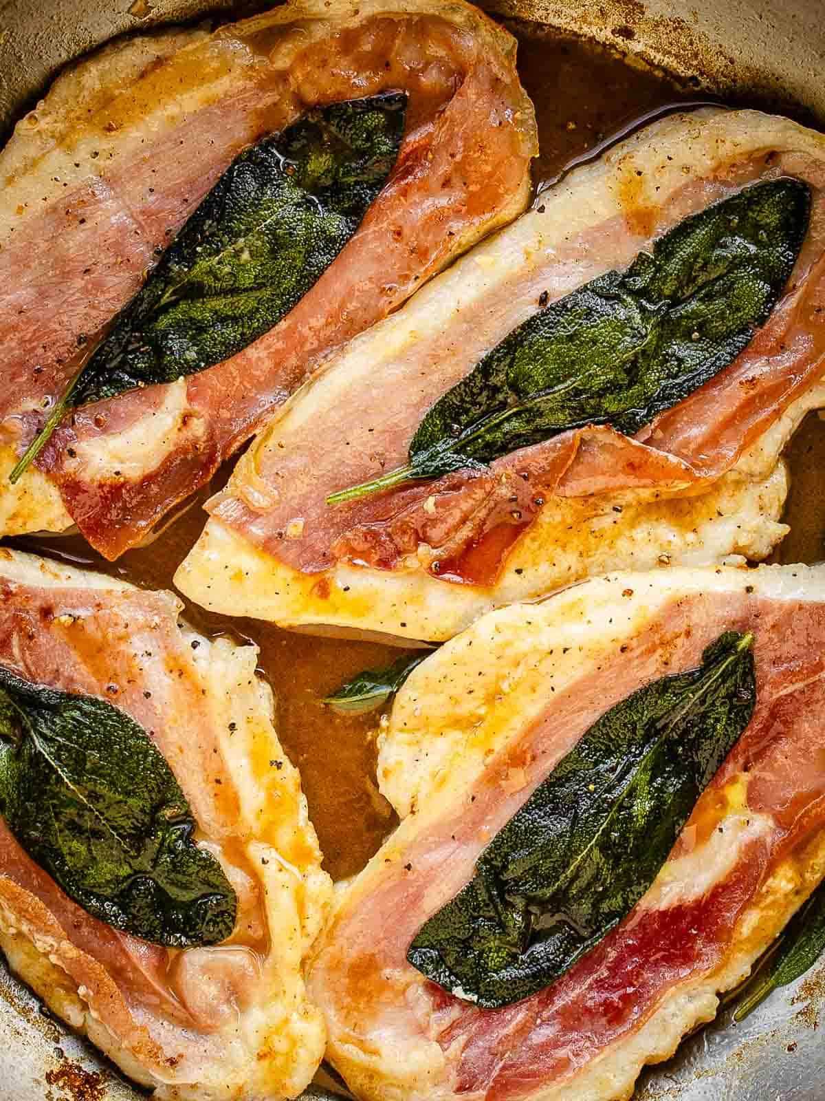 Four chicken cutlets with prosciutto and a sage leaf on each in a pan viewed from above.