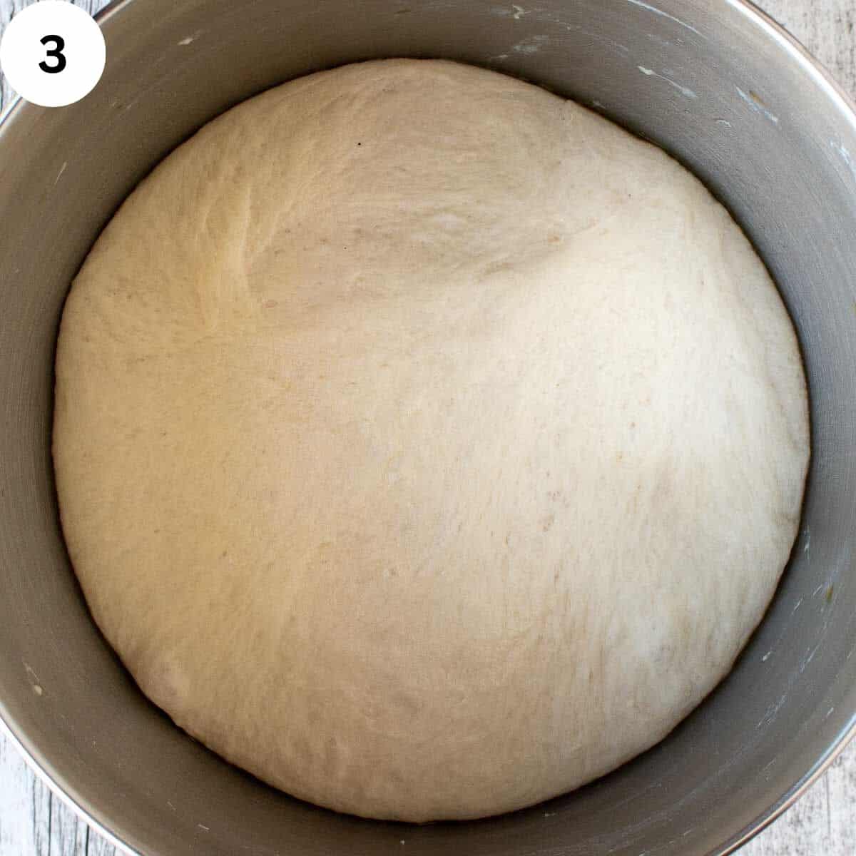 Risen dough in a metal mixing bowl after proofing, showing a smooth, airy surface.