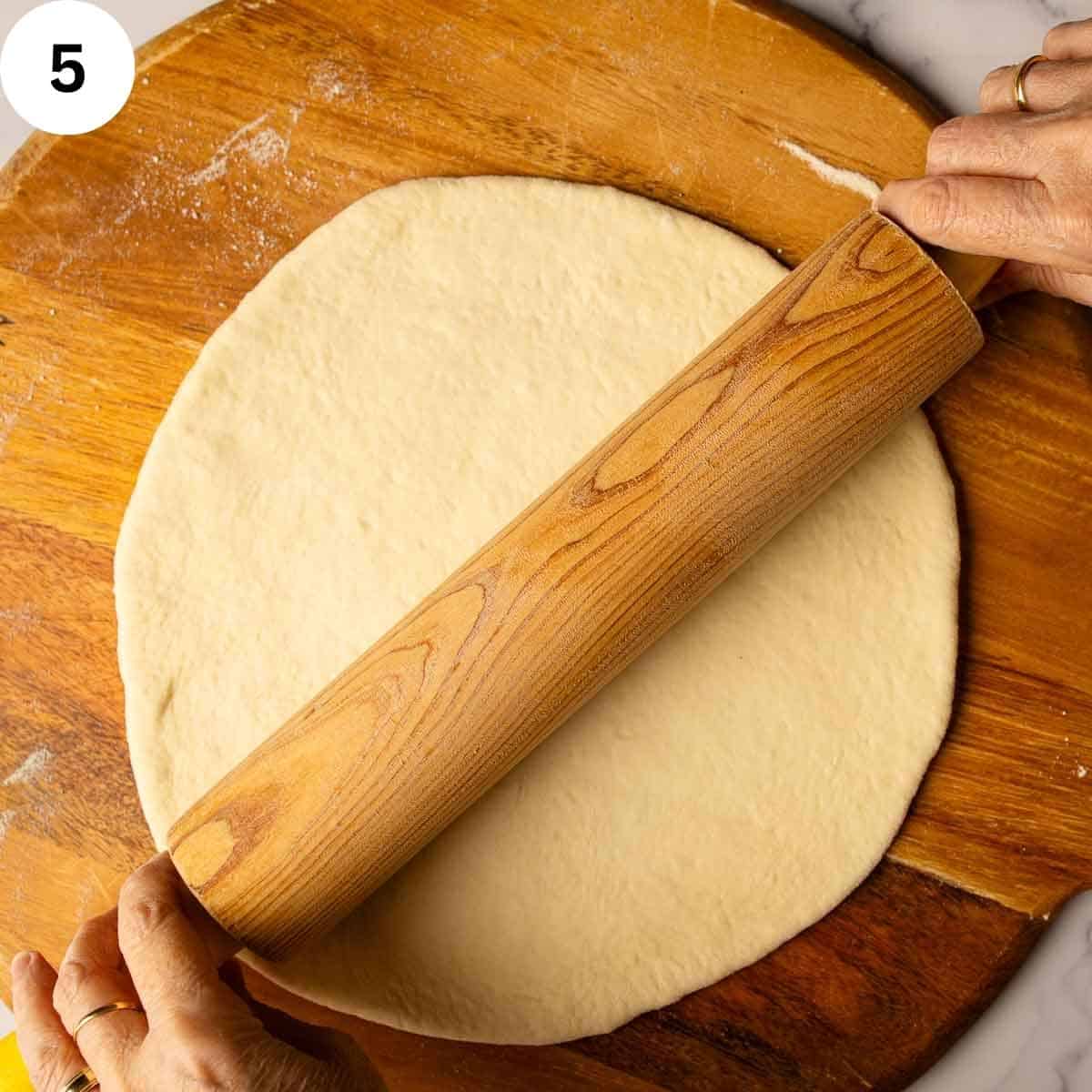 Rolling out dough into a round shape on a wooden board using a rolling pin.