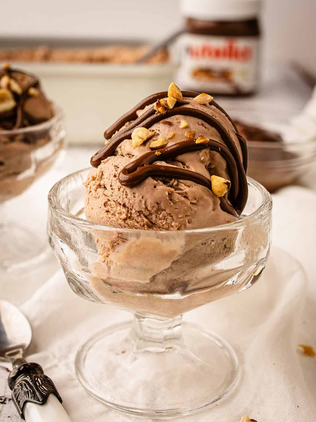 Nutella Ice Cream recipe scooped into dessert glass topped with a drizzle of nutella and chopped hazelnuts.