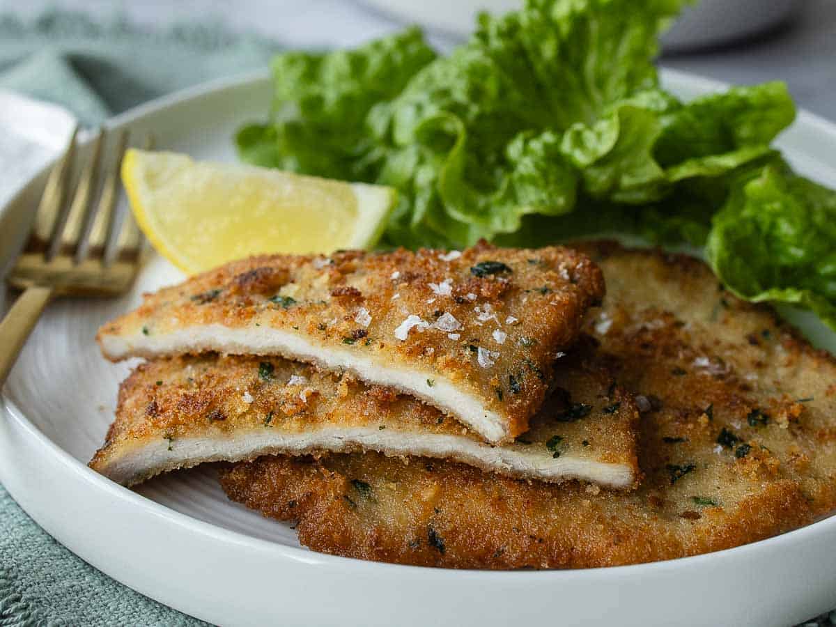 Cutlets on a plate, one cut in half to reveal the juicy meat, a wedge of lemon and green salad in the background.