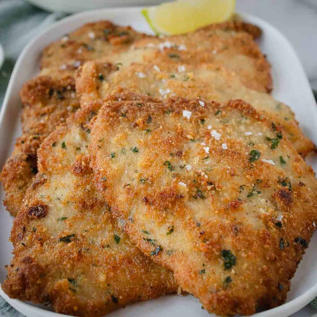 Pork cutlets on a oblong platter, sprinkled with flaked salt, a wedge of lemon and a salad in the background