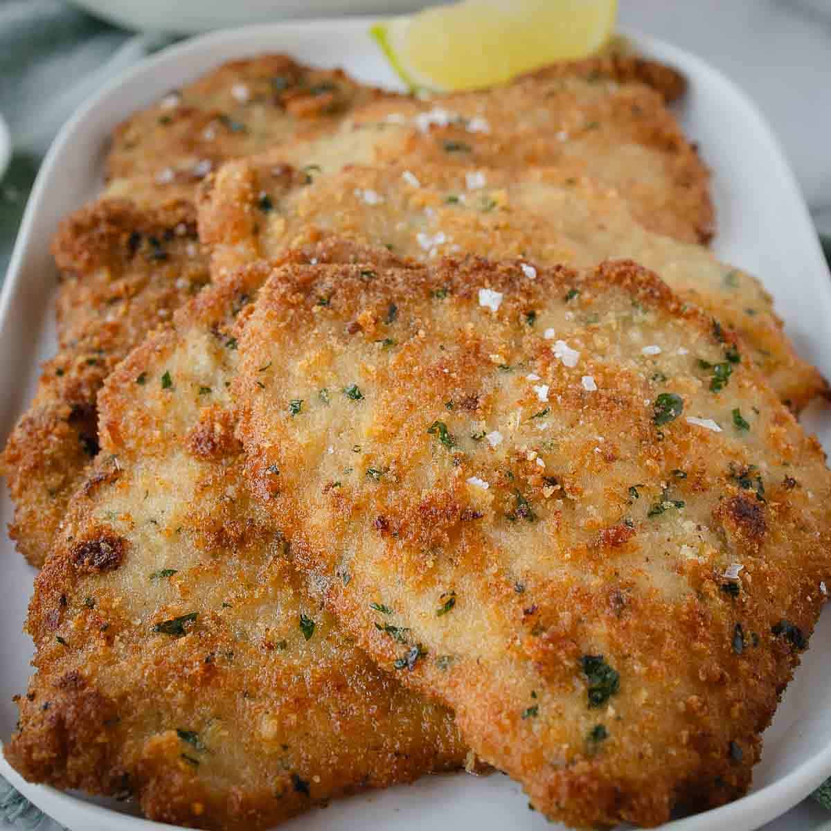 Pork cutlets on a oblong platter, sprinkled with flaked salt, a wedge of lemon and a salad in the background