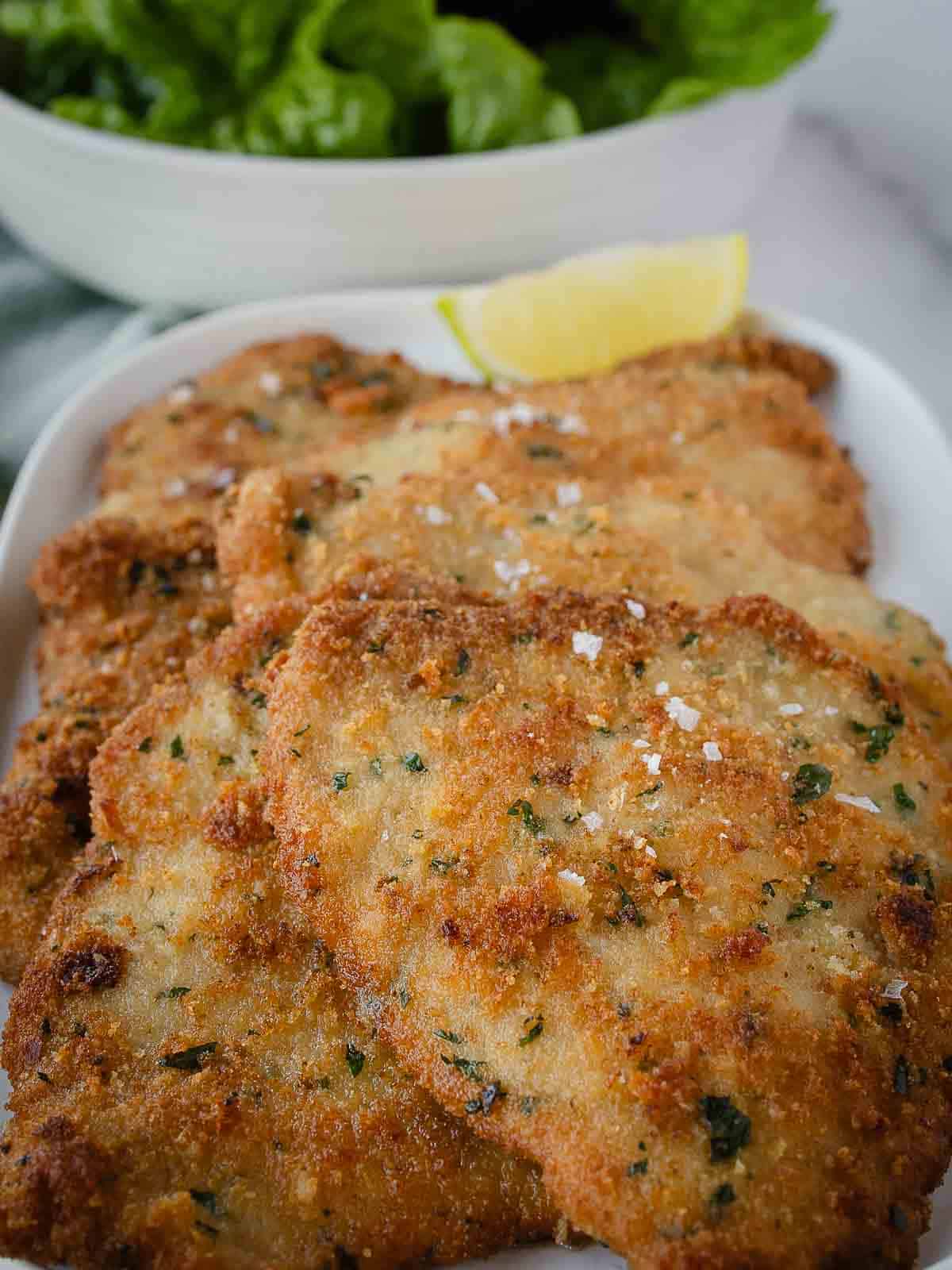 Pork cutlets on a oblong platter, sprinkled with flaked salt, a wedge of lemon and a salad in the background