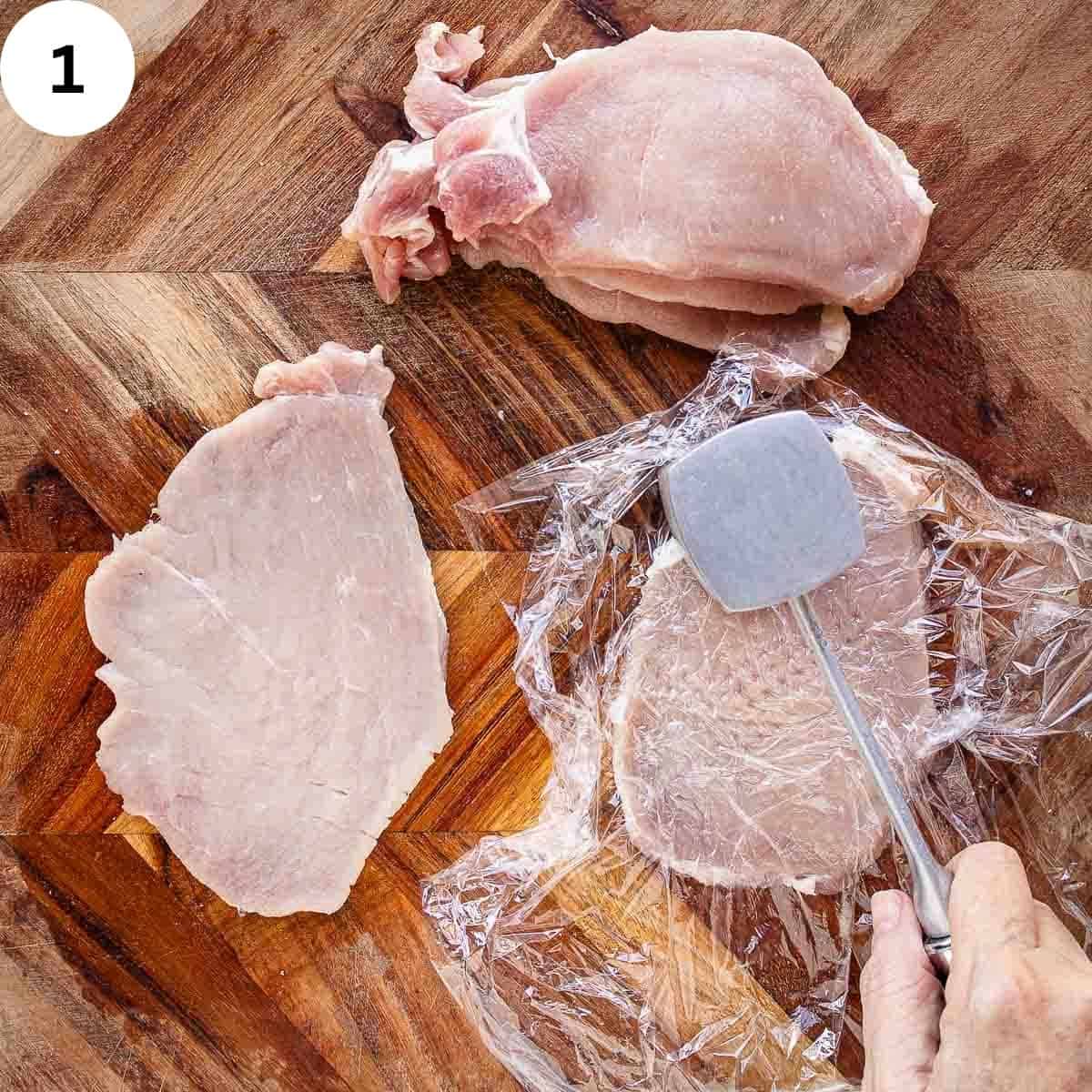 Slices of pork on a wooden board with one slice covered with plastic and being pounded with a meat mallet.