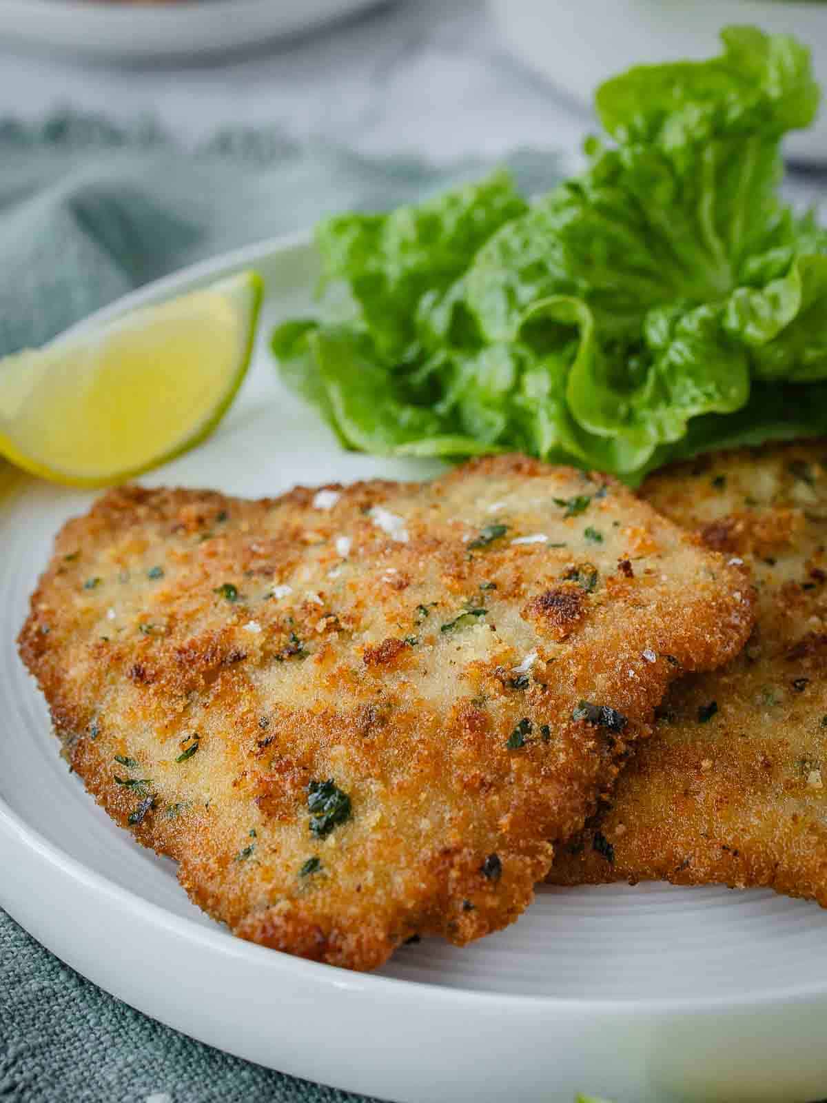 Close up of a breaded cutlet on a plate with a wedge of lemon and lettuce leaves.